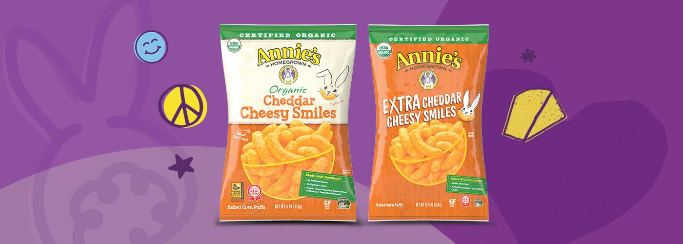 Single bags of Annie's Organic Cheddar Cheesy Smiles and Annie's Organic Extra Cheddar Cheesy Smiles Puffs on a purple background.
