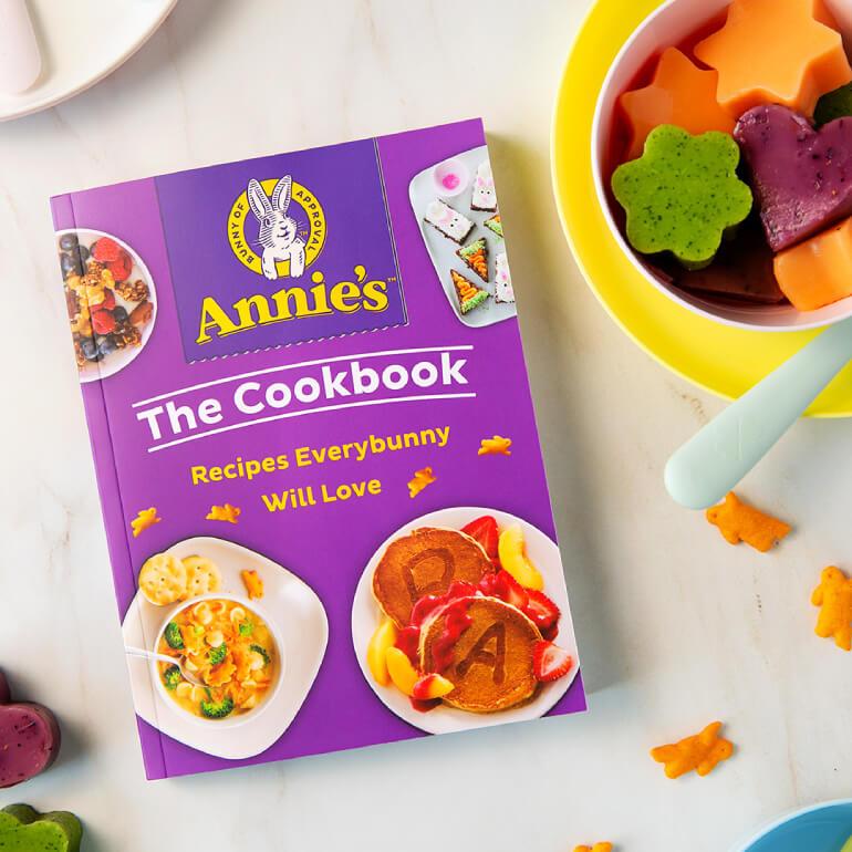 A book titled, "Annie's Cookbook, Recipes Every Bunny Will Love," sitting on a table with a bowl of fruit and some Cheddar Bunnies sprinkled around on a counter.