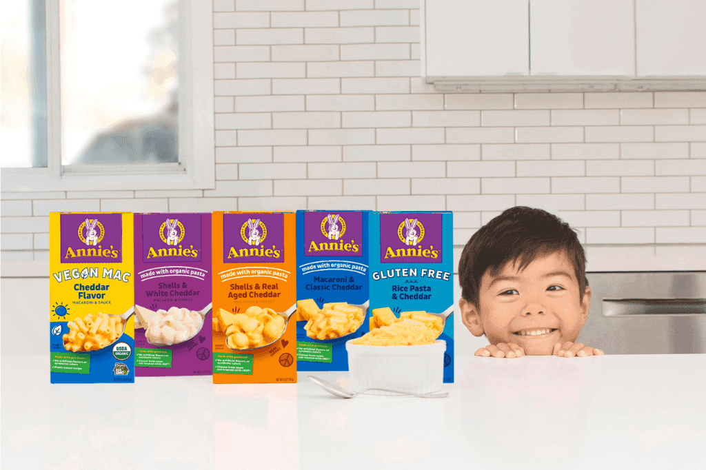 A smiling child peeks over a counter displaying five varieties of Annie's macaroni and cheese boxes in a kitchen.