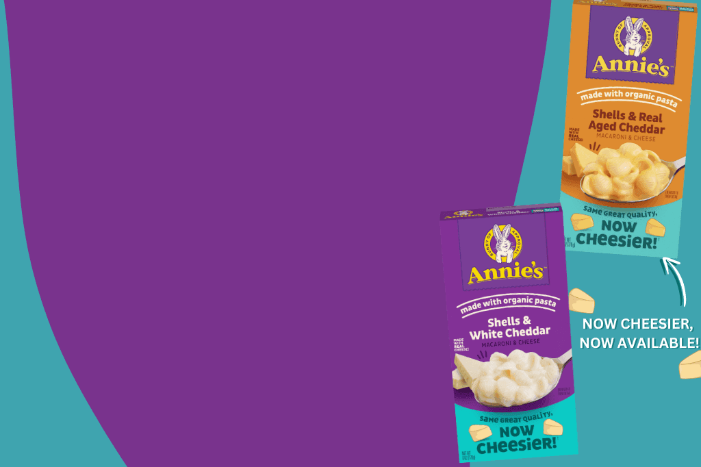 Two boxes of Annie's macaroni and cheese products on a vibrant purple and teal background. The left box is labeled 'Shells & White Cheddar,' and the right box is labeled 'Shells & Real Aged Cheddar.' Both boxes have the Annie's logo with a bunny mascot. The text on the bottom of the boxes says, 'NOW CHEESIER, NOW AVAILABLE!' with a white arrow