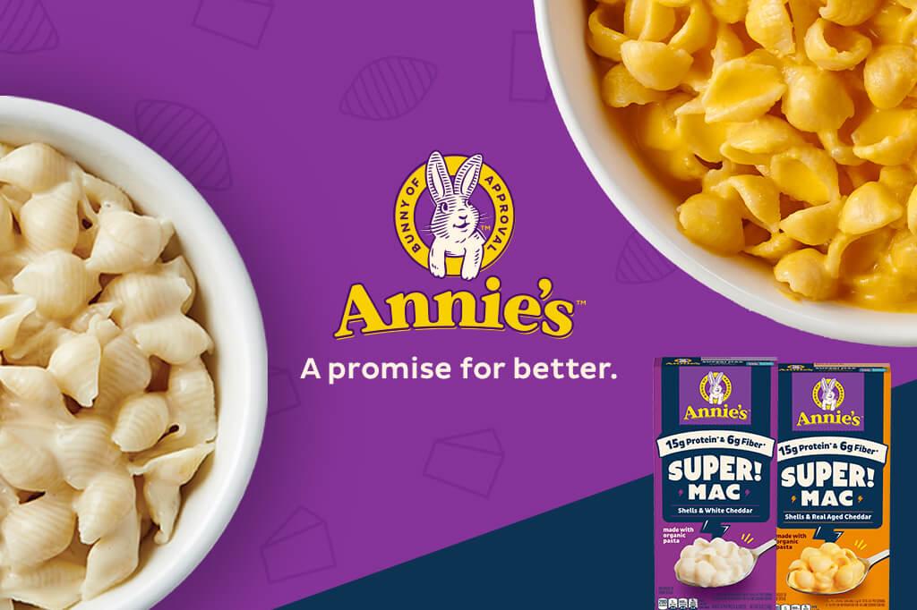 Two front facing pack shots of Super Mac Shells & White Cheddar and Shells Real-Aged Cheddar in the bottom right corner on a purple background with a bunny logo, two white bowls with Shells Mac in them and text in the middle has the Annies logo that says, "Annies's A promise for better."