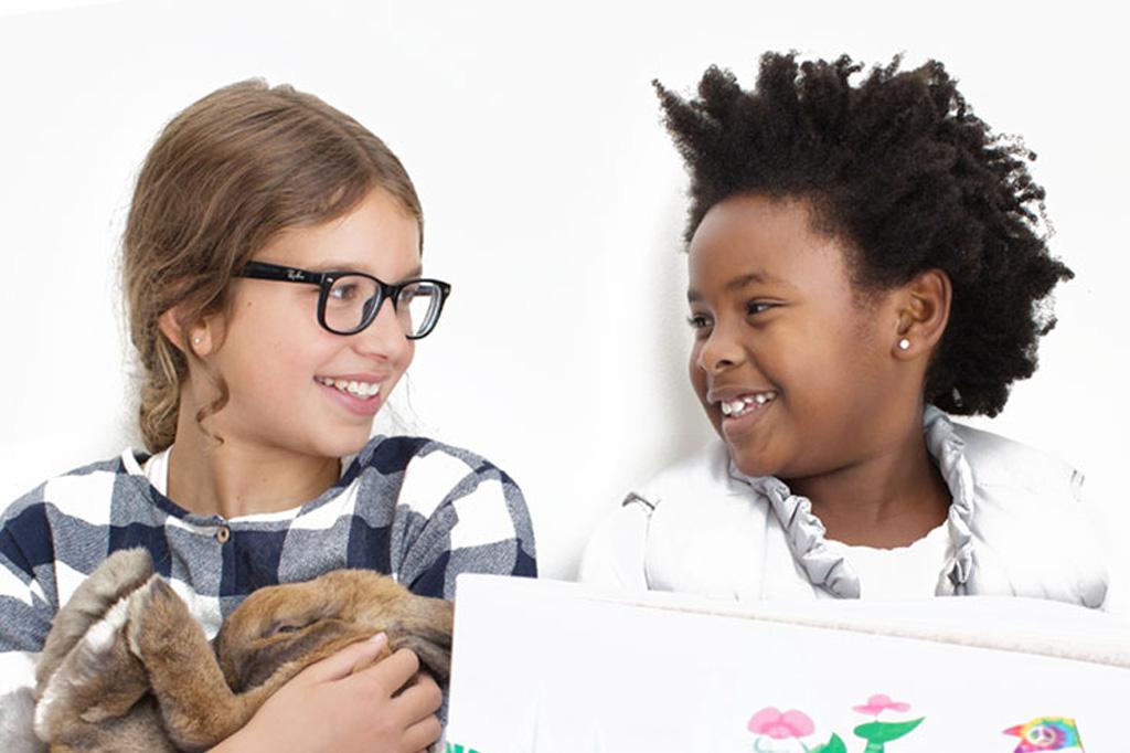 Two children smiling at each other, one holding a real bunny and the other holding a book.