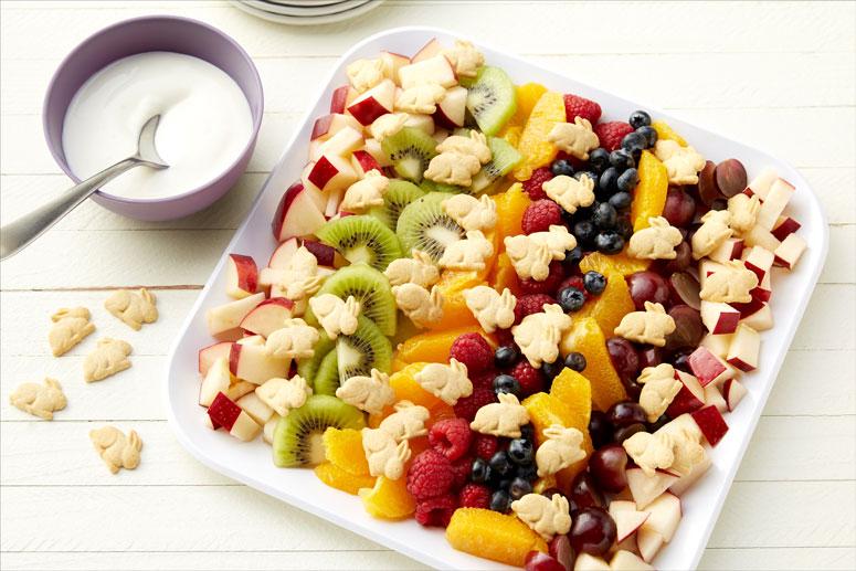 Bunny Fruit Salad on a square white plate with a small side bowl of vanilla yogurt and a few Annie's Organic Honey Bunny Grahams Baked Graham Snacks sprinkled on a surface.