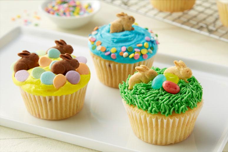 A white rectangle platter with three Bunny Graham Cupcakes frosted in different colors and sprinkled with Annie's Bunny Grahams, sprinkles and jelly beans.