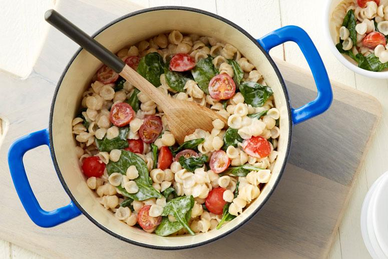 Overhead view of Spinach and Tomato Shells & Cheese in a large crock with a wooden spoon and sitting on a wood surface.