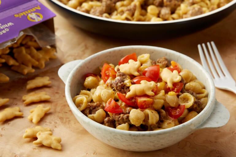 A bowl of an Annies recipe Cheeseburger Mac & Cheese with a white fork and an opened box with spilled Annie's crackers on the table