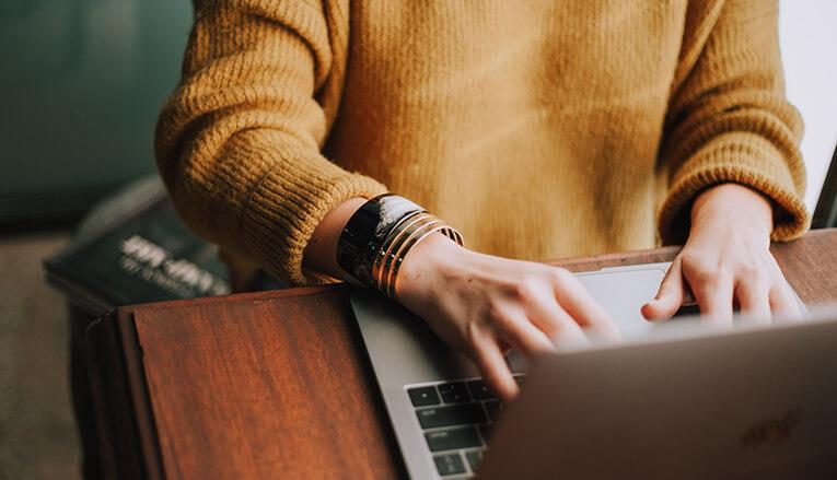 A close up image of someone typing on a laptop wearing an orange long sleeve shirt.