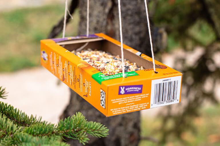 An empty Annie's box with strings attached and bird food in the box hanging from a tree.