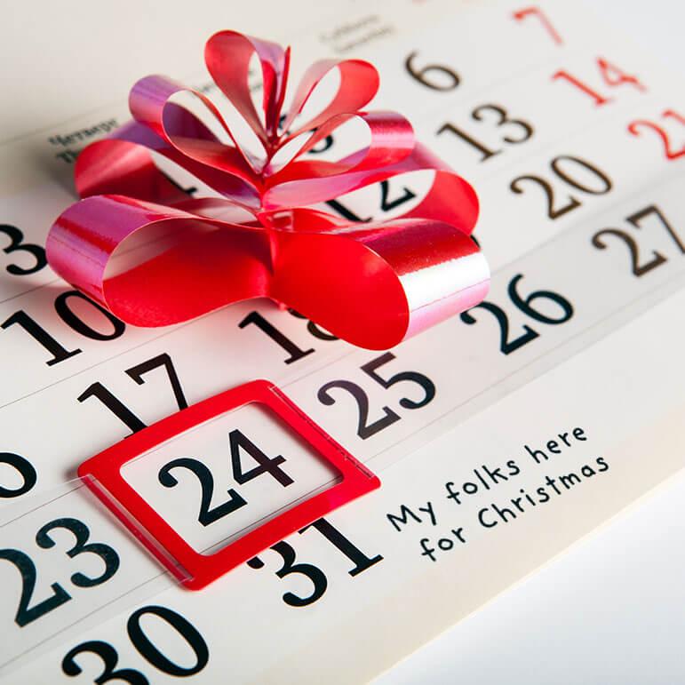 A close up of a calendar featuring a bright red bow and a red box around the 24th date with copy reading "My folks here for Christmas"