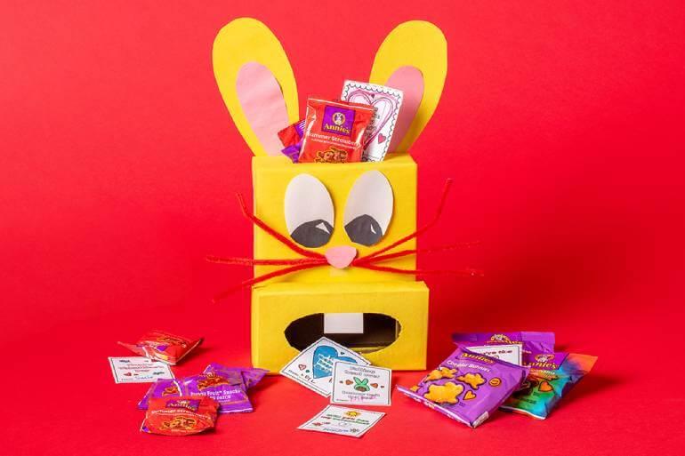 A yellow bunny box filled with Annie's snacks & activity cut outs in front on a red background